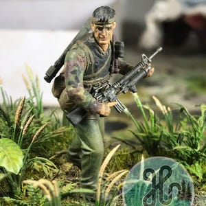 Könnte beinhalten: Eine handbemalte Miniatur-Soldatenfigur in Tarnkleidung und mit einem dunklen Stirnband, die ein schwarzes Gewehr hält. Die Figur ist inmitten von künstlichem Blattwerk und Gras positioniert, mit einem kreisförmigen Logo mit dem Text H DIORAMA.