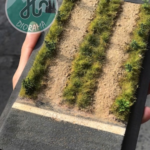 Pode incluir: Diorama em miniatura com uma estrada com uma linha branca, um caminho de terra e áreas de relva verde e pequenos arbustos. O diorama está sobre uma base preta. O logótipo "DIORAMA" é visível.