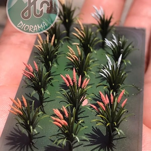 May include: A rectangular green base featuring twelve miniature grass tufts arranged in rows. Each tuft has dark green blades and a vertical flowering spike in shades of light pink, tan, or white. A circular logo with the text H DIORAMA is visible.