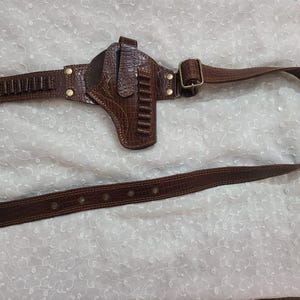 Op de afbeelding: Een bruine leren pistoolholster en riemset. De holster is ontworpen om een ​​handvuurwapen vast te houden en is bevestigd aan een riem met kogel lussen. De riem heeft een gesp en is gemaakt van hetzelfde bruine leermateriaal.
