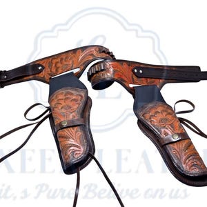 Op de afbeelding: Een bruine leren pistoolriem en holster set. De set heeft bloemmotieven en donkerbruine accenten. De holsters zijn zwart en de riem heeft een gesp. De set is ontworpen voor een Western thema.