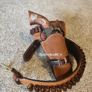 Puede incluir: Un juego de funda de cuero marrón con un revólver, cinturón y bucles de munición. El revólver tiene un mango decorativo. El cinturón tiene una hebilla de latón y está estampado con el texto "ZauberkraftCA".
