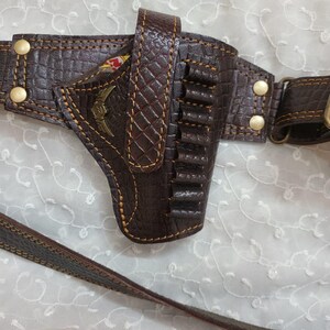 Op de afbeelding: Een bruin leren pistoolholster met een riem, met een textuurpatroon en decoratieve stiksels. Het holster heeft een compartiment voor een pistool en verschillende lussen voor kogels. Goudkleurige klinknagels en een decoratief embleem zijn zichtbaar.