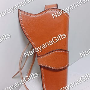 May include: A tan leather holster featuring a curved top edge, a horizontal strap across the middle, and visible white stitching along the seams. The holster includes a leather cord attached to the side and is marked with the text NarayanaGifts.