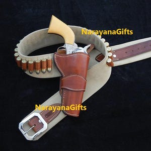 May include: A western-style gun belt with a brown leather holster, a silver revolver, and a tan suede belt with bullet loops. The belt has a silver buckle and a brown leather strap.