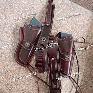 May include: A dark brown leather gun belt set featuring two holsters and a cartridge belt. The holsters and belt are adorned with silver-toned circular concho accents featuring a cross design. The belt includes loops for ammunition.