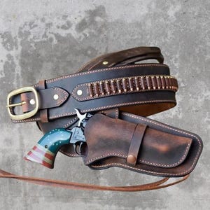 Puede incluir: Un cinturón de cuero marrón con una funda a juego y un revólver. El cinturón tiene una hebilla de latón y presillas para balas. La empuñadura del revólver está pintada con un diseño de bandera estadounidense.