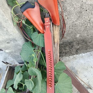Op de afbeelding: Een set van twee bruine leren pistoolholsters met zwarte toppen en een bijpassende bruine leren riem. De holsters en riem worden getoond tegen een rustieke houten achtergrond met groen gebladerte.