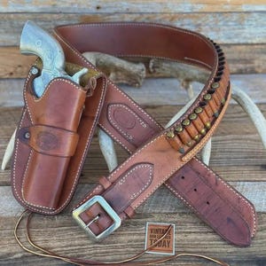 May include: A brown leather gun belt with a matching holster containing a silver-coloured revolver. The belt features a series of brass-cased cartridges held in loops, a metal buckle, and a small square tag labelled VINTAGE GUN LEATHER TODAY.