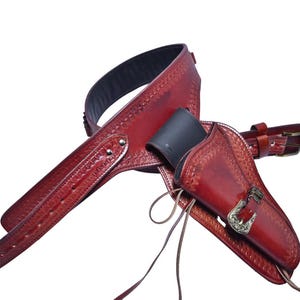 Op de afbeelding: Een rode leren pistoolriem en holster set. De riem is breed met decoratieve stiksels en een zwart interieur. De holster heeft een decoratieve gesp en is ontworpen om een pistool te bevatten. De set is gemaakt van leer.