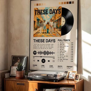 May include: A framed wall poster featuring a vinyl record graphic, the title These Days, a central illustration of five people walking down a street, a Spotify code, and a tracklist. The poster includes color swatches at the top and text details.