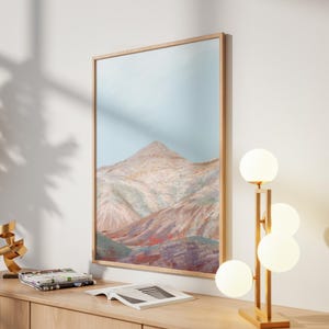 May include: A framed landscape art print depicts a mountain range in shades of beige, pink, and red under a light blue sky. A modern gold lamp with four spherical lights and a gold sculpture are on a wooden cabinet.