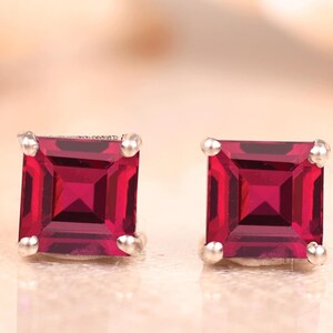 Puede incluir: Un par de pendientes de botón con piedras preciosas de color rojo intenso de corte cuadrado montadas en metal plateado. Cada pendiente presenta una piedra facetada con un engaste de cuatro garras, mostrando un tono carmesí vibrante.