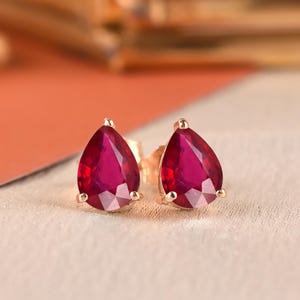 May include: A pair of stud earrings featuring pear-shaped, faceted deep pink stones set in gold-toned metal prongs. The earrings are positioned side-by-side, showcasing the vibrant color and the pointed teardrop silhouette of the gemstones.