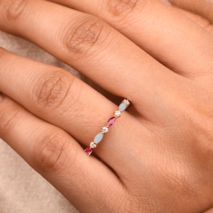 Könnte beinhalten: Ein dünner Goldring mit einem abwechselnden Muster aus rosafarbenen und hellblauen Steinen im Marquise-Schliff sowie kleinen runden, klaren Steinen, die in der oberen Hälfte des Metallrings gefasst sind und ein zartes Schmuckstück bilden.