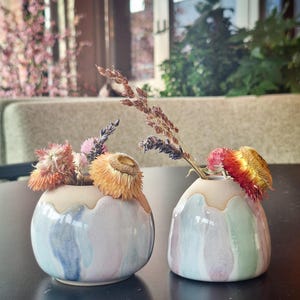 Op de afbeelding: Twee kleine, ronde keramische vazen met gedroogde bloemen. De vazen hebben een lichtblauw, roze en groen kleurenschema met een beige rand. Eén vaas heeft een blauw en roze aquareleffect. De andere vaas heeft een groen en roze aquareleffect.