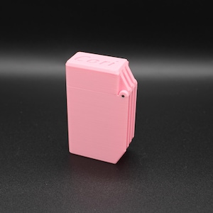 Puede incluir: Un encendedor rectangular rosa con la palabra "Cori" impresa en la parte superior. El encendedor tiene una tapa con bisagras y un dise&ntilde;o acanalado en un lado. El fondo es gris oscuro.