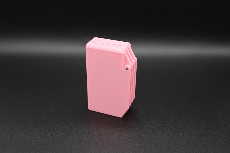 Puede incluir: Un encendedor rectangular rosa con la palabra "Cori" impresa en la parte superior. El encendedor tiene una tapa con bisagras y un dise&ntilde;o acanalado en un lado. El fondo es gris oscuro.