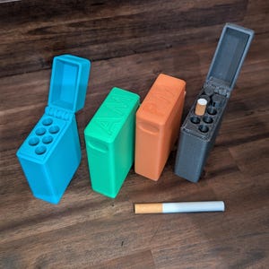 Puede incluir: Cuatro coloridos estuches para cigarrillos impresos en 3D en azul, verde, naranja y gris, con las letras "AM" y "PM" en dos de los estuches. Un cigarrillo es visible. Los estuches est&aacute;n sobre una superficie de madera.