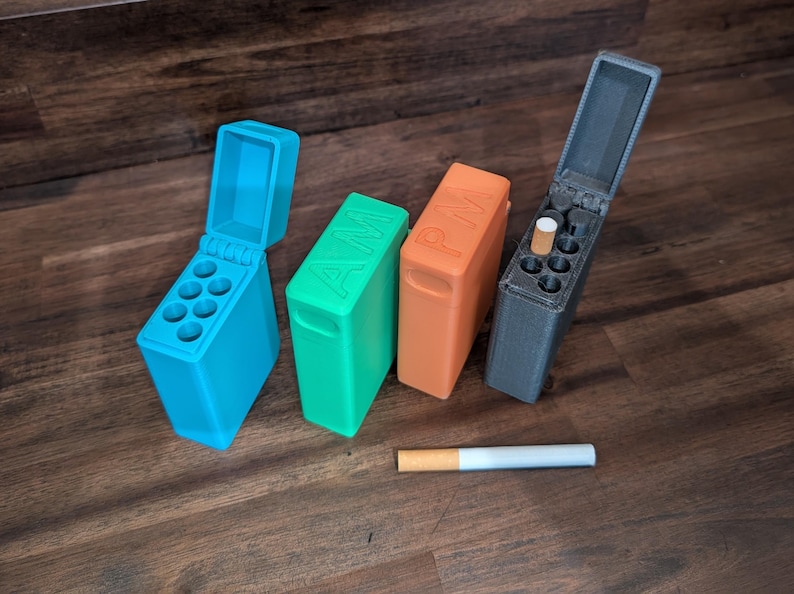 Puede incluir: Cuatro coloridos estuches para cigarrillos impresos en 3D en azul, verde, naranja y gris, con las letras "AM" y "PM" en dos de los estuches. Un cigarrillo es visible. Los estuches est&aacute;n sobre una superficie de madera.