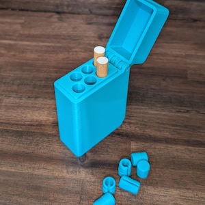 Puede incluir: Un estuche de cigarrillos turquesa impreso en 3D con una tapa abierta, que contiene dos cigarrillos. El estuche tiene seis compartimentos circulares y varias piezas cil&iacute;ndricas turquesas a juego est&aacute;n dispersas cerca. El estuche est&aacute; sobre una superficie de madera oscura.