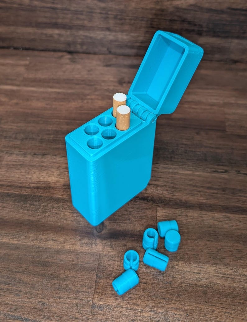 Puede incluir: Un estuche de cigarrillos turquesa impreso en 3D con una tapa abierta, que contiene dos cigarrillos. El estuche tiene seis compartimentos circulares y varias piezas cil&iacute;ndricas turquesas a juego est&aacute;n dispersas cerca. El estuche est&aacute; sobre una superficie de madera oscura.
