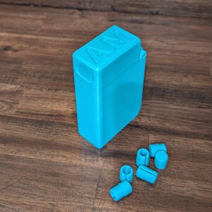 Puede incluir: Un contenedor rectangular turquesa brillante con las letras "AM" en relieve en la tapa. Varias piezas cil&iacute;ndricas turquesas peque&ntilde;as est&aacute;n dispersas cerca. Los objetos est&aacute;n sobre una superficie de madera.