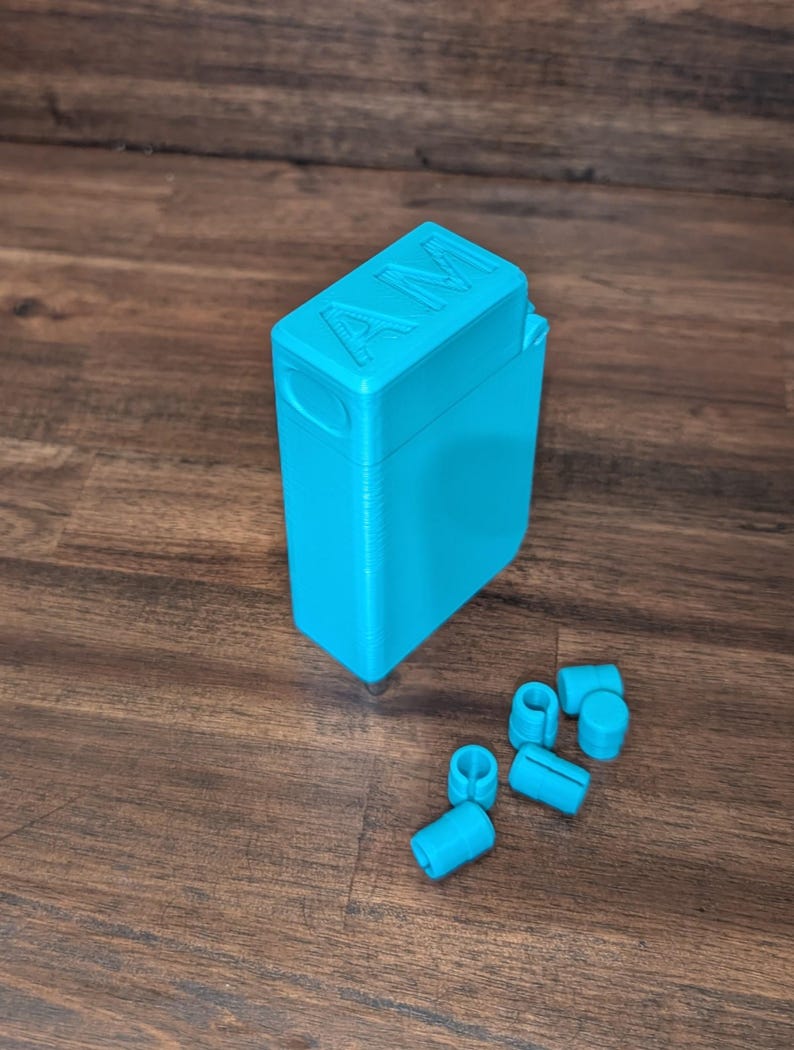 Puede incluir: Un contenedor rectangular turquesa brillante con las letras "AM" en relieve en la tapa. Varias piezas cil&iacute;ndricas turquesas peque&ntilde;as est&aacute;n dispersas cerca. Los objetos est&aacute;n sobre una superficie de madera.