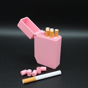 Puede incluir: Un pitillera rosa abierta, que revela varios cigarrillos. Varios filtros rosas y un cigarrillo est&aacute;n junto a la pitillera. El fondo es negro.