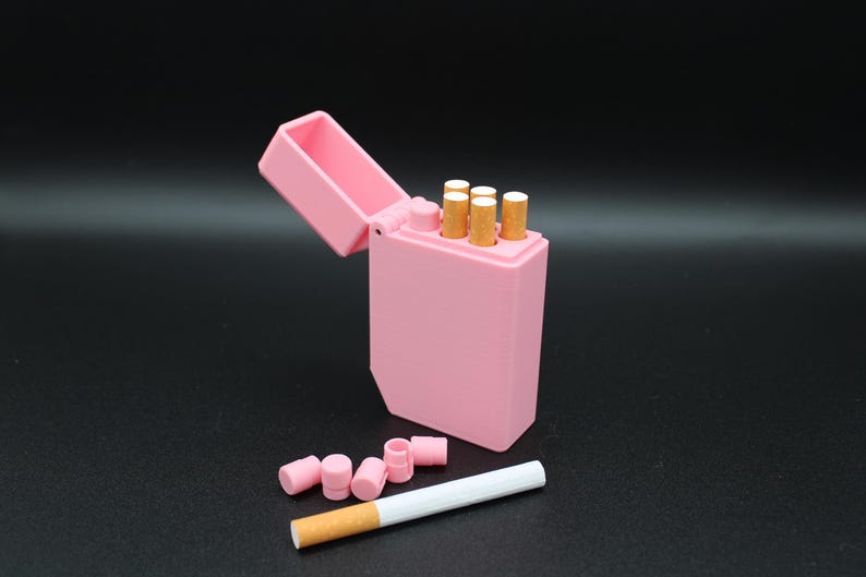 Puede incluir: Un pitillera rosa abierta, que revela varios cigarrillos. Varios filtros rosas y un cigarrillo est&aacute;n junto a la pitillera. El fondo es negro.