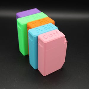 Puede incluir: Una fila de estuches rectangulares de colores en tonos morado, verde, naranja, azul y rosa. Los estuches tienen una tapa con bisagras y texto en la parte superior, incluyendo "PM", "AM" y "Cori".