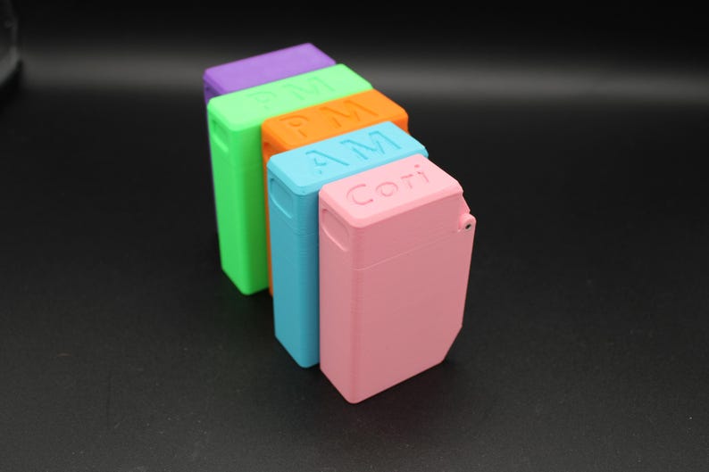 Puede incluir: Una fila de estuches rectangulares de colores en tonos morado, verde, naranja, azul y rosa. Los estuches tienen una tapa con bisagras y texto en la parte superior, incluyendo "PM", "AM" y "Cori".