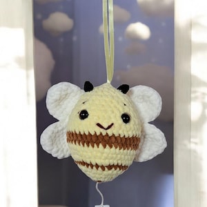 May include: A handmade crochet bee mobile with a yellow body, brown stripes, and white wings. The bee has black eyes and a smiling mouth. A white plastic ring hangs below the bee, and a green ribbon is attached to the top.