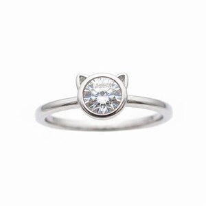 May include: A silver ring with a round, clear gemstone set in a bezel. The bezel is shaped like a cat's face, with small ear-like protrusions. The band is thin and polished, creating a minimalist design.