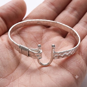 May include: A silver-colored metal bangle bracelet featuring a hammered, textured surface. The bracelet has a hook-and-eye style clasp mechanism with a coiled wire detail wrapping around one side of the band near the opening of the hook.