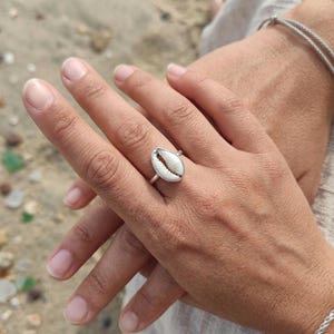 May include: A silver ring with a white and tan seashell design. The ring's band is silver, and the seashell has a natural shape. The ring is worn on a hand, with a beach background visible.