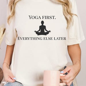 Könnte beinhalten: Cremefarbenes T-Shirt mit dem Text "YOGA FIRST. EVERYTHING ELSE LATER" und einer schwarzen Silhouette einer Person in einer Yoga-Pose. Die Person hält einen rosa Becher. Die Person trägt zerrissene blaue Jeans.