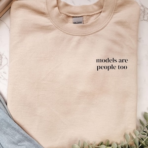 Könnte beinhalten: Beiges Sweatshirt mit Rundhalsausschnitt und dem schwarzen Text "models are people too". Das Sweatshirt liegt gefaltet auf einer Marmoroberfläche, mit blauen Jeans und Grünzeug im Hintergrund.