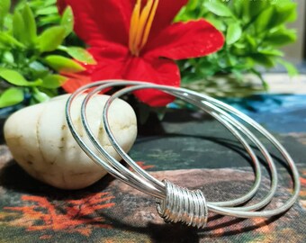 Peut inclure: Trois bracelets argentés avec un motif enroulé au centre. Les bracelets sont présentés devant une pierre blanche, une fleur rouge et un feuillage vert. L'image est posée sur un fond coloré à motifs.