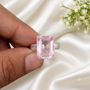 May include: A silver ring featuring a large, rectangular, faceted light pink gemstone set in a simple metal bezel. The stone has a clear, transparent appearance with visible internal facets that reflect light, mounted on a smooth, polished silver band.