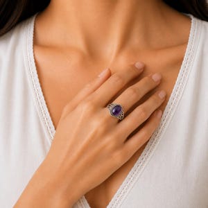 May include: A silver ring featuring an oval-shaped purple amethyst gemstone set in a decorative metal band with twisted rope detailing and intricate scrollwork patterns along the sides of the setting, worn on a finger.
