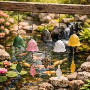 May include: A collection of colorful wind chimes hanging from a wooden beam. The chimes are bell-shaped and come in green, pink, black, white, and yellow. Each chime has a decorative leaf-shaped clapper. The background features a pond and lush greenery.