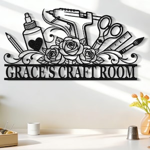 Puede incluir: Letrero de metal negro con el texto "GRACE'S CRAFT ROOM" y herramientas de artesanía decorativas, incluyendo tijeras y una pistola de pegamento. El letrero está montado en una pared blanca sobre una mesa con suministros de artesanía.