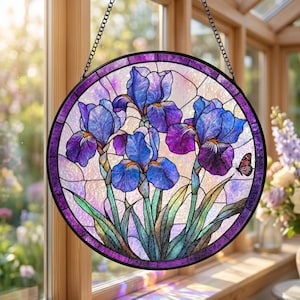 Puede incluir: Vidriera redonda con flores de iris azules y moradas y una pequeña mariposa. La obra de arte está enmarcada en un borde de metal negro y cuelga de una cadena. El fondo es de color rosa claro y blanco.