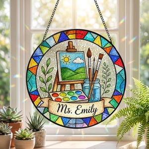 May include: A round, stained-glass window art piece with a colorful geometric border. The central design features a painting scene with an easel, brushes, and a palette. A banner at the bottom reads "Ms. Emily". The piece hangs from a chain.