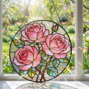 Puede incluir: Vidriera redonda que representa tres rosas rosas con hojas y tallos verdes. La obra de arte está enmarcada en negro y cuelga de una cadena. Se ve un jardín soleado en el fondo.