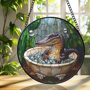 May include: A circular stained glass window depicting an alligator in a bathtub with bubbles. The alligator has a brown and purple body. The background features green plants and a blue sky. The window is suspended by a black metal chain.
