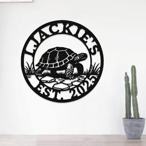 May include: Black metal wall art featuring a turtle design inside a circular frame. The text "JACKIE'S" is at the top, and "EST. 2023" is at the bottom. The turtle is on a stone path with grass.