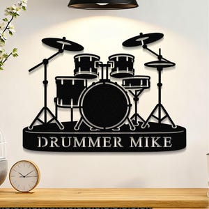 以下が含まれることがあります： 「DRUMMER MIKE」の文字が入った黒い金属製のドラムセットの壁掛けアート。 ドラムセットには、バスドラム、スネアドラム、シンバル、スタンドが含まれています。 音楽愛好家のための装飾品。