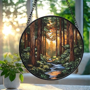 Puede incluir: Una ventana redonda de vidrieras que representa una escena de bosque con árboles altos, un arroyo y luz solar. La obra de arte presenta tonos de marrón, verde y naranja, encerrada en un marco de metal negro y suspendida por una cadena.